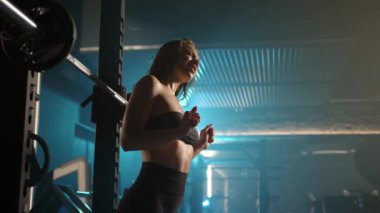 Neşeli genç bir kadın spor salonundaki halter setleri arasında neşeli molalar verir, dans eder ve şarkı söyler, halter halterine enerji katar. Kamera 8K HAM. 