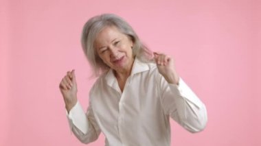 Gri saçlı neşeli yaşlı bir kadın pembe bir arka planda beyaz bir bluzla neşeyle dans ediyor, pozitiflik ve hayattan keyif alıyor. Kamera 8K HAM. 