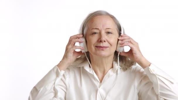一位快乐的老妇人 戴着耳机 一边听着令人振奋的音乐 一边笑着 穿着经典的白色衬衫 体现着快乐和音乐上的愉悦 8K照相机 — 图库视频影像