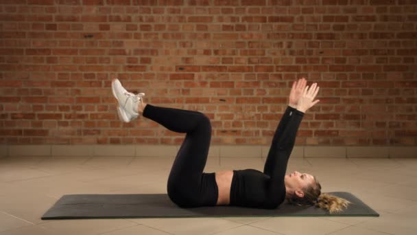 一名妇女在健身房的垫子上与她的核心肌肉接触 做左边的死虫子运动 专注于控制运动 以提高砖墙前的稳定性和腹部力量 照相机前置 — 图库视频影像