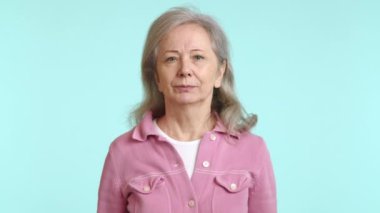 Mor gömlekli yaşlı bir kadın yüzünde mutlu bir gülümsemeyle öpücük savurarak bir jest yapıyor. Bir etkinlikte eğleniyor ve eğleniyor. Kollarını ve sakallarını gösteriyor.