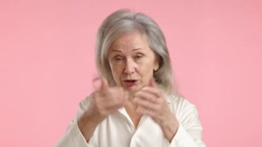 Yaşlı bir kadın, yumuşak pembe bir arka planı gerilimin altını çizen bir videoda kesin bir şekilde işaret ettiği için güçlü bir hoşnutsuzluk ve memnuniyetsizliği ifade ediyor. Yüksek kalite 4k görüntü