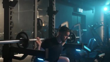 Spor salonundaki zinde bir adam halter çömelmelerini güvenli bir şekilde gerçekleştirir. Çağdaş bir fitness ortamında vücudunun alt kısmını güçlendirir. Kamera 8K HAM. 