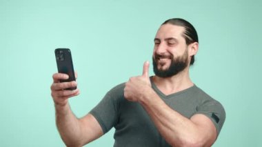 Uzun saçlı neşeli bir adam bir selfie çeker, telefonunu kaldırırken geniş bir tebessüm eder, açık deniz mavisi arka planında eğlenceli bir anı yakalar. Kamera 8K HAM.