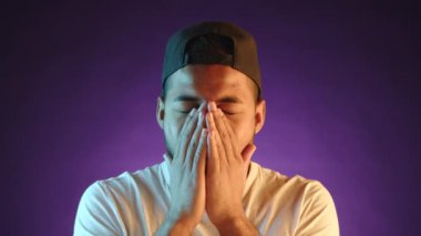 Sıradan bir atmosferde, melez bir erkek, mor bir arka planda dinlenmeye ihtiyacı olduğunu göstermek için ağzını eliyle kapatarak kendini uyuşuk hisseder ve esner. Kamera 8K HAM. 