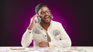 Diş teli takan neşeli Afro-Amerikalı erkek girişimci mor bir arka planda nakit parayla çevrili bir telefon yığını kullanıyor. Kamera 8K HAM.