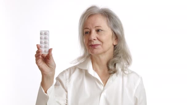 灰色の髪を持つ高齢女性は 薬のブリスターパックを保持し 疑念と懐疑を示しています 白い背景に隔離された白いブラウスを身に着けています 医療遵守の概念に最適です カメラ Raw — ストック動画
