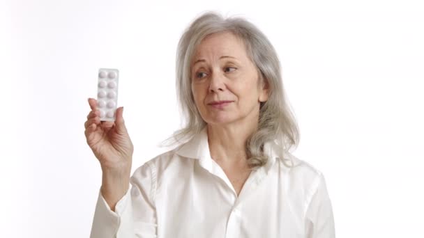 熟考的な表現を持つ高齢女性は 彼女の健康オプションと白い背景での潜在的な治療を考慮して 丸薬のブリスターパックを保持しています カメラ Raw — ストック動画