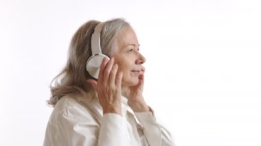 Yaşlı bir kadın kulaklık takıyor, kendini sakin müziğin huzurlu melodisine daldırıyor, dinlenme ve müzik terapisinin tadını beyaz bir arka planda çıkarıyor. Kamera 8K HAM. 