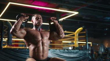 Konsantre olmuş erkek vücut geliştirici, güçlü kas yapısını vurgulamak için canlı spor salonu ışıklarıyla çevrili kettlebell kaldırma hareketleri yapıyor. Kamera 8K HAM. 