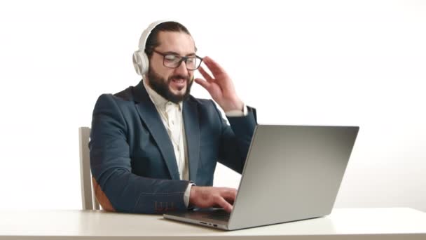 長い暗いひげとメガネを持つ幸せなビジネスマンは 現代の仕事の柔軟性と満足の明確な表現で彼のラップトップを使用しながら ヘッドフォンを使用して 彼の仕事で楽しみを見つける — ストック動画