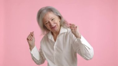 Gri saçlı neşeli yaşlı bir kadın pembe bir arka planda beyaz bir bluzla neşeyle dans ediyor, pozitiflik ve hayattan keyif alıyor. Kamera 8K HAM. 