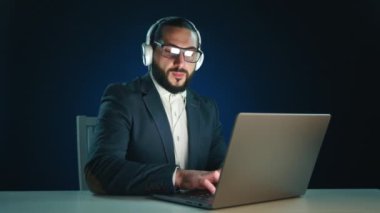 Koyu renk takım elbiseli ve kulaklıklı profesyonel bir adam koyu mavi arka planda dizüstü bilgisayar üzerinde çalışırken müziğe dalmış. İş kavramlarında çoklu görev ve rahatlama için mükemmel bir görsel