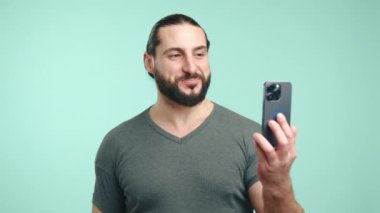 Sakallı neşeli bir iş adamı neşeli bir video görüşmesi için akıllı telefonunu kullanıyor, sakin bir deniz mavisi arka planında gülümsüyor ve hareketsiz bir şekilde konuşuyor. Kamera 8K HAM.