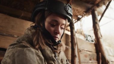 Kaskı ve taktik giysisi olan odaklanmış bir kadın asker ahşap bir sığınağın içinde, zihinsel olarak yakın bir harekete hazırlanıyor. Kamera 8K HAM. 