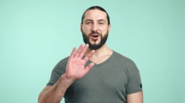 Uzun saçlı ve günlük gri tişörtlü dost canlısı bir işadamı, elini geniş bir gülümsemeyle sallayarak, deniz mavisi arka planda bir misafirperverlik ve yakınlık hissi uyandırıyor. Kamera 8K HAM.