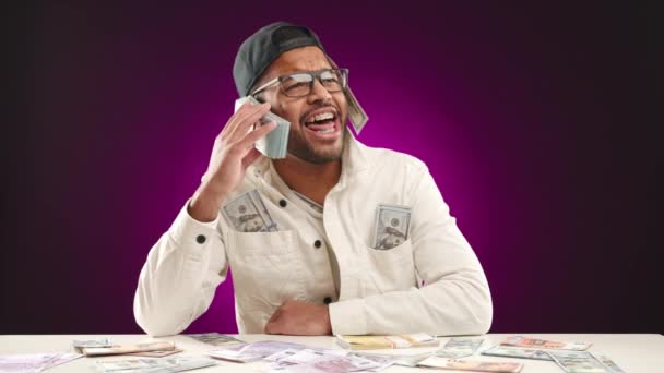 一个兴高采烈的人坐在一张满是钱的桌子旁 沉浸在幽默的繁荣的景象中 一边玩一边用一叠美元钞票进行电话交谈 象征着财富和成功 — 图库视频影像