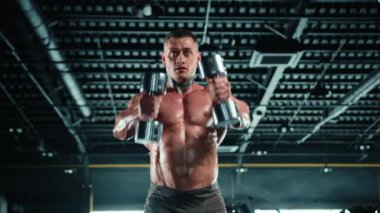 Terli, kaslı bir adam spor salonunda yüksek kontrastlı ışıklandırmayla ağır dambılları rahatça kaldırır. Onun yoğun bakışları ve fiziği güç yayıyor. Kamera 8K HAM. 