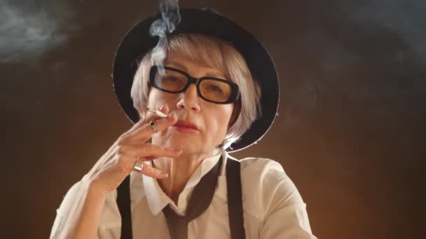 神秘的老妇人自信地在一个褐色背景的黑暗房间里抽烟 烟充满了她周围的空间 她穿着黑社会风格的衣服 戴着吊带衫 戴着一顶黑色的帽子 — 图库视频影像