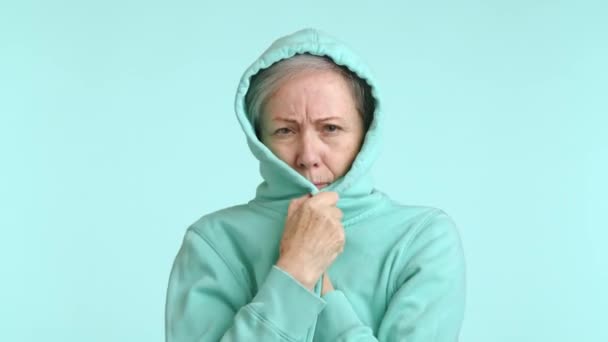 高齢の女性は柔らかいフードに身を包み 冷たい色の背景に対して暖かさと快適さを求めているように見えます カメラ Raw — ストック動画