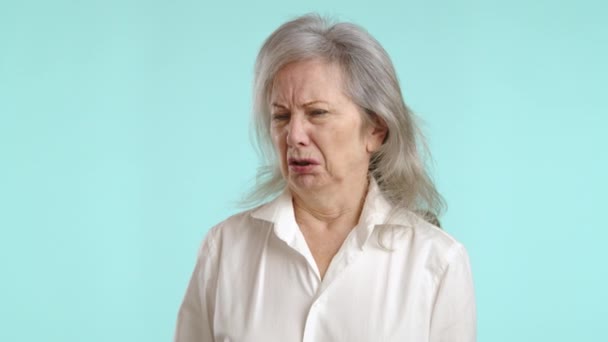 不快な年配の女性は鼻をピンチし 彼女の顔は嫌悪感に震え 強い不快な臭いを示す対照的なターコイズの背景に立っています カメラ Raw — ストック動画