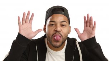 Neşeli bir Afro-Amerikalı adam, saf beyaz bir arka planda kaygısız ve neşeli bir titreşim yayarak ellerini kaldırarak, neşeli bir şekilde dilini uzatır. Kamera 8K HAM. 