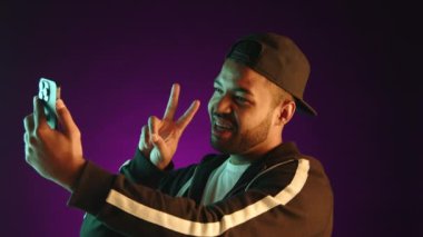 Canlı bir genç adam bir eliyle barış işareti gösterirken diğer eliyle ustalıkla selfie çekiyor, yüzü dinamik mor bir arka plana karşı neşeyle parlıyor. Kamera 8K HAM. 