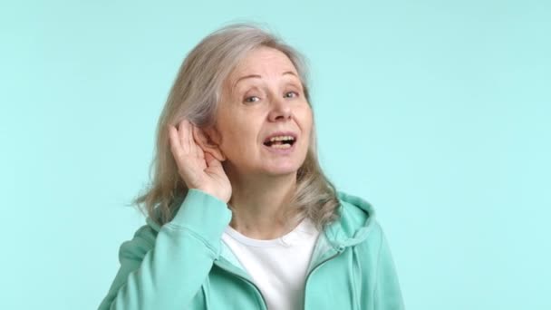 注意深い年配の女性は彼女の耳に彼女の手を保持し 遠くの会話や騒音を聞くために緊張し 静かなシアン背景に対して 音を拾うために集中した努力を表現します — ストック動画