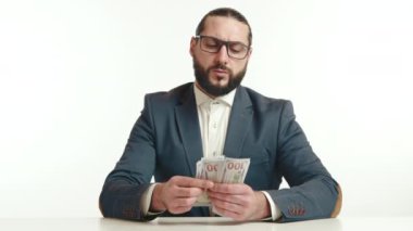 Mavi takım elbiseli ve beyaz gömlekli odaklanmış bir iş adamı finansal analiz ve para yönetimine odaklanmış bir yığın doları dikkatle sayıyor. Kamera 8K HAM.