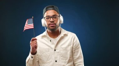 Diş teli takan neşeli bir çift ırklı adam en sevdiği melodileri sever. Koyu mavi arka planda gururla Amerikan bayraklarını sallar. Kamera 8K HAM. 