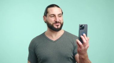 Sakallı neşeli bir iş adamı neşeli bir video görüşmesi için akıllı telefonunu kullanıyor, sakin bir deniz mavisi arka planında gülümsüyor ve hareketsiz bir şekilde konuşuyor. Kamera 8K HAM.