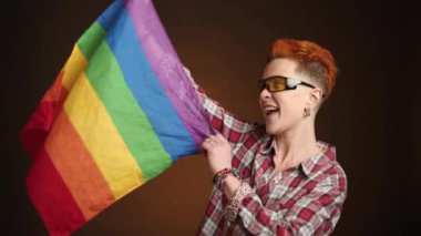 Mutluluk saçan, şık güneş gözlüklü lezbiyen bir kadın LGBT bayrağını kaldırıyor, kendinden geçmiş ifadesi karanlık bir arkaplanda özgürlüğün ve seçimin sembolü. Kamera 8K HAM.