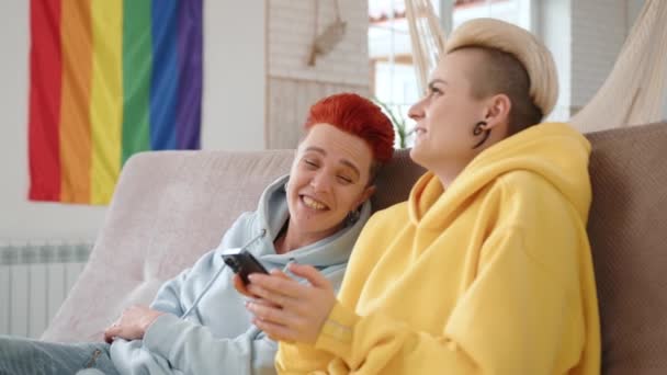 两位女同性恋在家里的沙发上分享着一个舒适而快乐的时刻 她们沉浸在智能手机的内容中 旁边挂着一面象征男女同性恋 双性恋和变性者的旗帜 高质量的4K镜头 — 图库视频影像