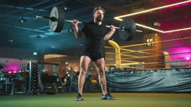 Yoğunlaşmış bir erkek atlet halter çömelmeleri, duruşu ve şekli spor salonunun dinamik ışıklandırmasındaki çalışmanın yoğunluğunun altını çiziyor. Kamera 8K HAM. 