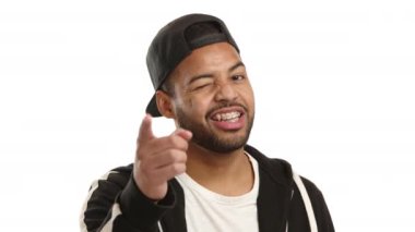 Oyuncu bir melez adam göz kırpıyor ve kameraya bakıyor, diş teli takıyor ve siyah bir şapka takıyor, beyaz bir arka planda ortak bir sırrı paylaşıyormuş hissi veriyor. Kamera 8K HAM. 