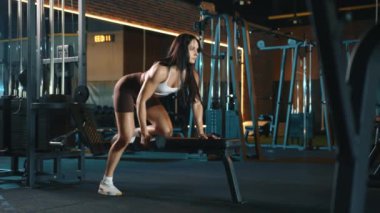 Şortlu bir kadın spor salonundaki bir bankta dambılı kaldırıyor ve sırt kasları üzerinde çalışıyor. Spor aletleri ve döşemeleri olan bir binada eğlence etkinliği düzenleniyor.