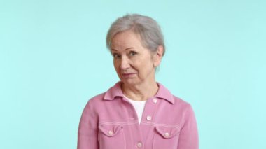 Pembe gömlekli, sıcak bir gülümsemesi olan yaşlı bir kadın, rahatlatıcı, mavi bir arka plana karşı güven ve pozitiflik yayıyor ve onayını başını sallayarak gösteriyor. Kamera 8K HAM. 