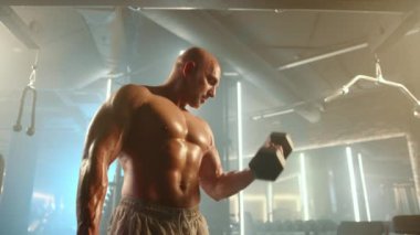 Kafası tıraşlı kaslı bir adam, iki elinde de dambıllar olan pazularıyla yoğun bir şekilde çalışıyor, odaklandığı yer doğal ışıkla dolu bir spor salonu. Kamera 8K HAM. 