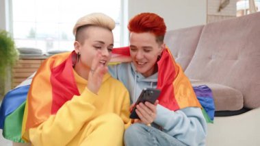 Gökkuşağı pelerinlerine sarılı genç bir lezbiyen çift, modern ve kapsayıcı bir aşkı sembolize eden akıllı bir telefona bakarken gülücükler ve neşe dolu bir an paylaşıyor. Kamera 8K HAM. 