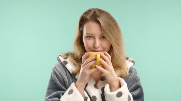 若い女性は穏やかな朝を楽しみ 穏やかなターコイズの背景に居心地の良いポルカドットローブに包まれている間 黄色いカップからお茶を飲む — ストック動画