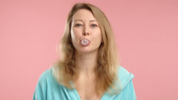 輝かしい瞬間に捉えられた 空の青いローブの若い女性がバブルガムを噛み パステルピンクの背景に大きな泡を吹き飛ばします — ストック動画