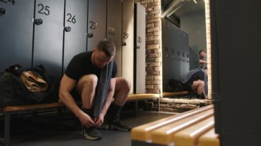 Spor salonundaki kendini işine adamış bir sporcu ayakkabı bağcıklarını dikkatle bağlıyor ve soyunma odasının sakin atmosferiyle çevrili sıkı bir antrenman sonrası tatminini yansıtıyor..