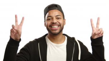 Dişlerinin üzerinde diş teli takan neşeli bir adam barış işaretini iki eliyle de sevinçle gösterir. Dostça tavırları beyaz bir arkaplanda vurgulanmıştır. Kamera 8K HAM. 