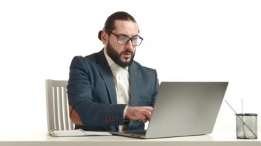 Takım elbiseli bir iş adamı beyaz bir masanın üzerindeki dizüstü bilgisayarın yanına notlar yazarak düzenli çalışma alışkanlıklarını sembolize ediyor. İş planlaması ve verimlilik konuları için ideal. Kamera 8K HAM. 