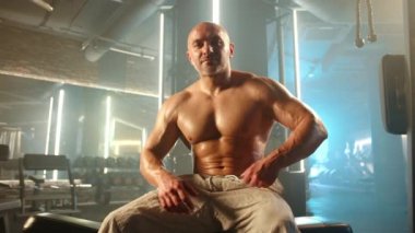 Çıplak vücutlu, kaslı bir vücut geliştirici spor salonundaki bir bankta oturuyor, kaslı kalçalarını ve karnını göstermek için şort giyiyor, güçlü bir göğüs ve tanımlanmış insan bacaklarıyla.