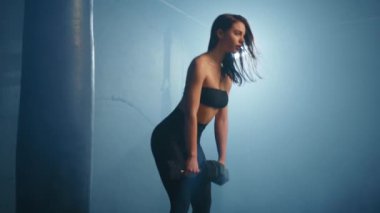 Bir fitness meraklısı, modern bir spor salonunun puslu atmosferinde canlı bir arka ışığa karşı silueti ile dambıllarla eğilir ve kararlılığını vurgular. Kamera 8K HAM.