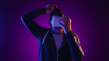 Hayal gücü yüksek ve dalgın bir adam interaktif bir sanal gerçeklik oyunu oynarken yüzünü canlı mor renkler sarmış bir şekilde VR kulaklığı ile kaplayan görünmeyen bir dünyaya uzanır. Kamera Ham.