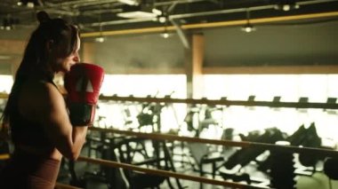 Antrenman kıyafetli bir kadın boksör, boks ringinde odaklanma ve kararlılıkla hava yumruk tekniği uyguluyor. Atmosferik bir spor salonunda dövüş senaryosu canlandırıyor. Kamera 8K HAM. 