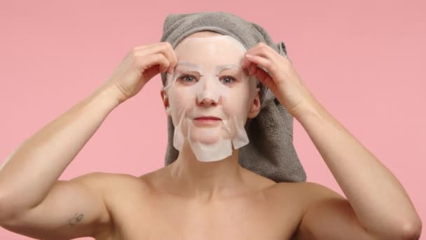 タオルターバンを着用している女性が 彼女のスキンケア体制を強調するために柔らかいピンク色の背景に繊細に水和する顔のマスクを適用する穏やかなシーン — ストック動画