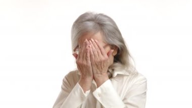 Detaylı bir şekilde yakalanan yaşlı bir kadının yüzü korku içinde donuyor. Kafasını düz beyaz bir zemine yaslıyor. Saf korku ve endişeyi resmediyor. Kamera 8K HAM. 